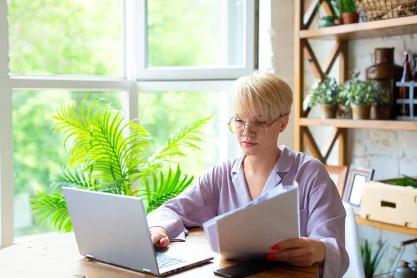 Γυναίκα στην πιτζάμα εργάζεται με έγγραφα μπροστά από ένα φορητό υπολογιστή οθόνη στο εσωτερικό του σπιτιού. Ελέγχει τα φορολογικά αρχεία πριν υποβάλει την αναφορά.. — Φωτογραφία Αρχείου