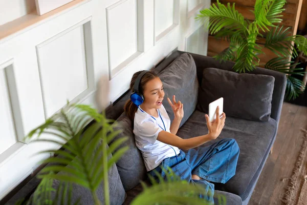 Teenager girl in blue headphones during online video call in home interior. Online meeting with friends.