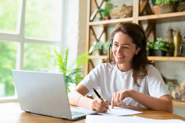 Uma mulher sorrindo trabalha com papéis em casa na frente de um monitor de laptop. Ela está verificando papéis durante a chamada de vídeo online. — Fotografia de Stock