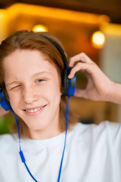 Foto vertical de um adolescente ruivo com fones de ouvido azuis. Ele ouve música e sorri. — Fotografia de Stock