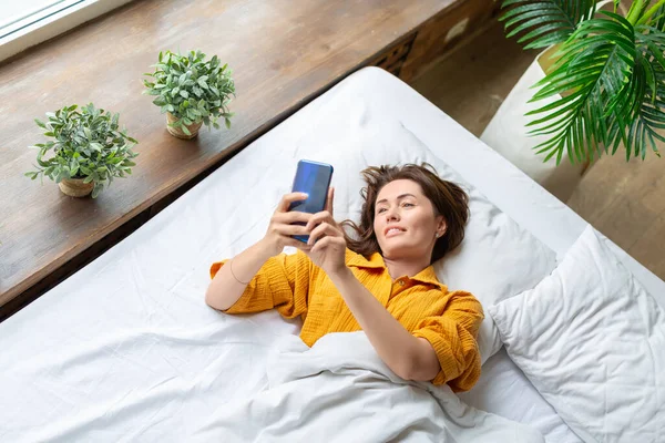 Φωτογραφία μιας γυναίκας στο κρεβάτι που ελέγχει τα μηνύματά του στο smartphone μετά την αφύπνιση. — Φωτογραφία Αρχείου