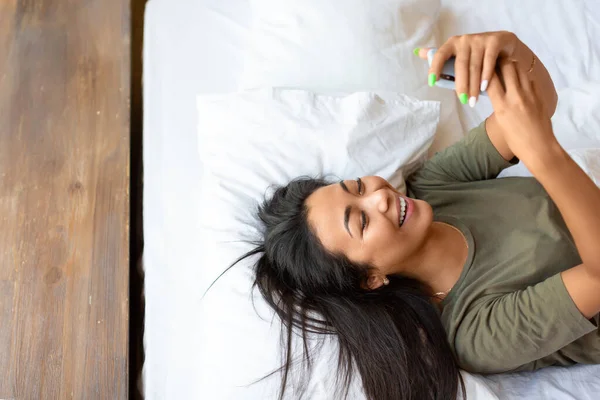 웃고 있는 행복 한 동양 여자 핸드폰을 들고 침대에 누워 있다. — 스톡 사진