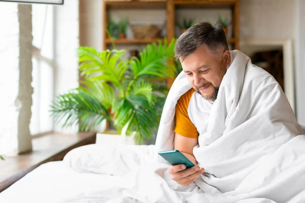 Pria paruh baya di tempat tidur memeriksa pesannya di smartphone setelah terbangun. Kecanduan terhadap konsep jejaring sosial. — Stok Foto