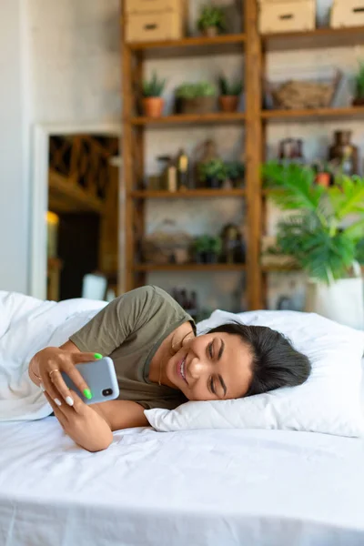 한 아시아인 여성의 수직 사진 이 휴대폰을 들고 침대에 누워 있습니다. 일어나서 소셜 미디어 게시물을 보고 있었어요. — 스톡 사진