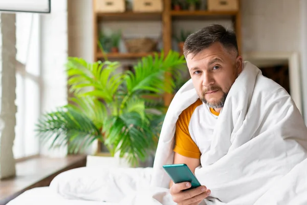 Seorang pria paruh baya yang sakit duduk di tempat tidur di bawah selimut. Dia memegang smartphone dan akan memanggil dokternya untuk konsultasi online. — Stok Foto