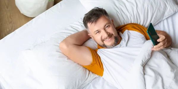 Smiling middle aged man with smartphone looking at camera. Dia berbaring di tempat tidur dengan smartphone dan memeriksa berita terbaru dari jejaring sosial. — Stok Foto