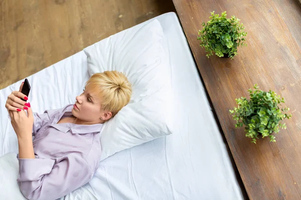 Vue de dessus d'une femme se trouve dans le lit avec smartphone. Elle vérifie les médias sociaux après le réveil. Concept de technologie moderne. — Photo