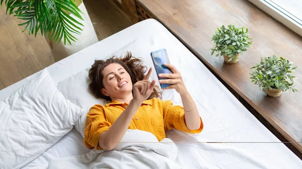 Μια χαμογελαστή γυναίκα βρίσκεται στο κρεβάτι με το κινητό τηλέφωνο μετά την αφύπνιση. — Φωτογραφία Αρχείου