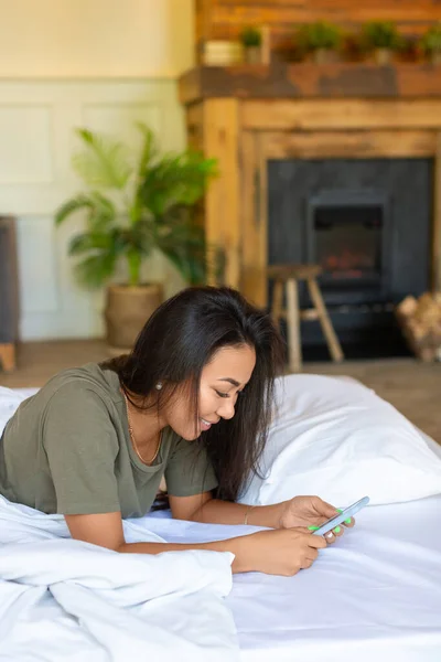 Foto vertikal dari wanita Asia yang bahagia tertawa terletak di tempat tidur dengan ponsel. Dia menerima pernyataan cinta dari pacarnya. — Stok Foto