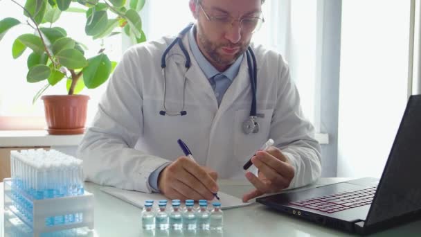 Ο γιατρός δουλεύει μπροστά από ένα φορητό υπολογιστή. Κρατάει ένα δοκιμαστικό σωλήνα με ένα δείγμα αίματος στο χέρι του και συντάσσει έκθεση για το έργο που επιτελείται στον τομέα της ανοσολογίας και του εμβολιασμού.. — Αρχείο Βίντεο