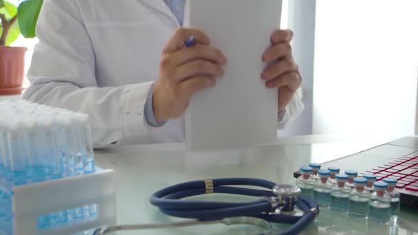 Dokter bekerja di depan monitor laptop. Dia menggesek kertas, meletakkan pena dan melepas kacamatanya. Sepertinya dia mendapat masalah penelitian. — Stok Video