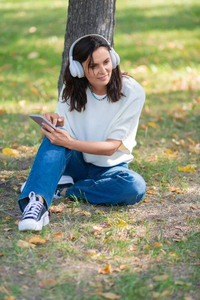 Photo d'une femme souriante de 40 ans dans un casque avec un smartphone. Elle écoute sa musique préférée tout en étant assise sur l'herbe dans le parc et prend une pause de l'agitation de la ville. — Photo