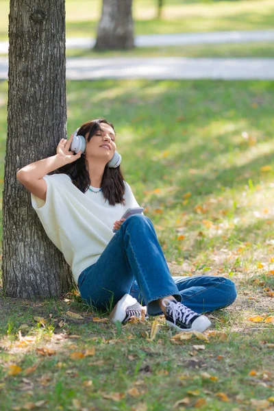 Femme de 40 ans portant un casque avec un smartphone. Elle écoute sa musique préférée tout en étant assise sur l'herbe dans le parc et prend une pause de l'agitation de la ville. — Photo