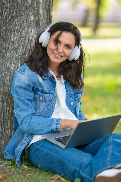 Femme d'âge moyen souriante avec ordinateur portable dans le parc de la ville. Concept de travail en ligne ou d'éducation en ligne. Photo verticale. — Photo