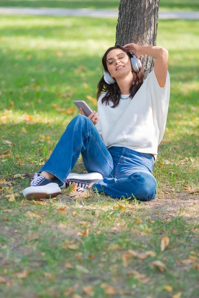 Zdjęcie 40-letniej kobiety w słuchawkach ze smartfonem. Słucha swojej ulubionej muzyki siedząc na trawie w parku i robi sobie przerwę od zgiełku miasta. — Zdjęcie stockowe