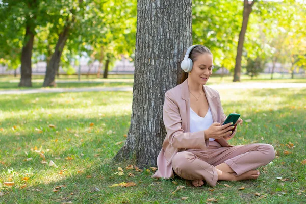 Photo d'une femme souriante de 40 ans dans un casque avec un smartphone. Elle écoute sa musique préférée tout en étant assise sur l'herbe dans le parc et prend une pause de l'agitation de la ville. — Photo