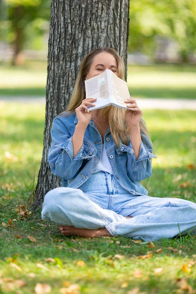 Pionowe zdjęcie kobiety z książką w parku. Siedzi na trawie i śmieje się, zakrywając twarz książką. — Zdjęcie stockowe