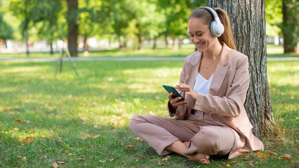 Photo d'une femme souriante de 40 ans dans un casque avec un smartphone. Elle écoute sa musique préférée tout en étant assise sur l'herbe dans le parc et prend une pause de l'agitation de la ville. — Photo