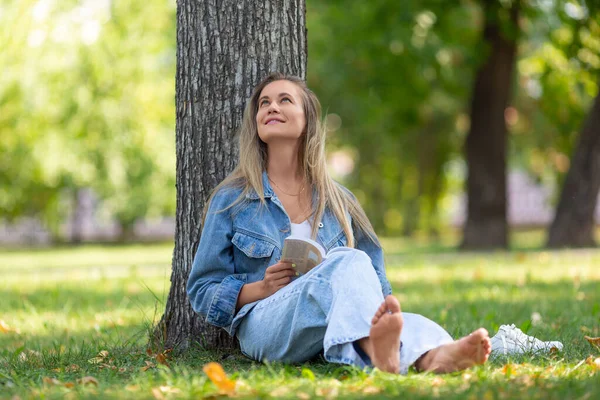 Szczęśliwa śmiejąca się kobieta z książką w parku. Siedzi na trawie i śmieje się - zabawna opowieść. — Zdjęcie stockowe