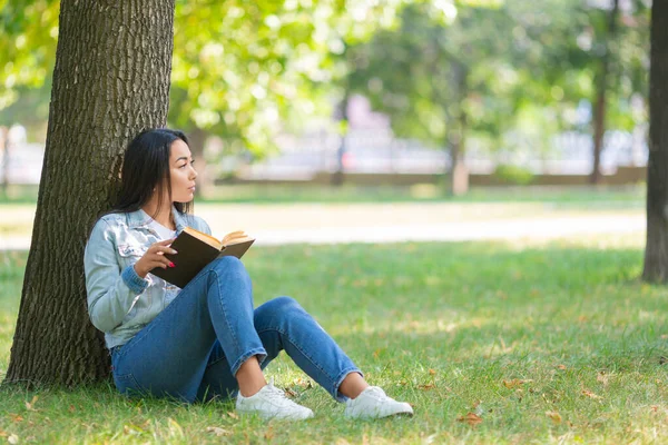 Azjatka z książką w parku. Siedzi na trawie w parku i patrzy obok siebie. Koncepcja detoksykacji cyfrowej. — Zdjęcie stockowe