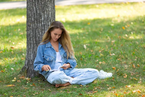 Uma mulher com um livro no parque. Ela se senta na grama no parque e lê uma história. O conceito de descanso de aparelhos, smartphones e Internet. — Fotografia de Stock