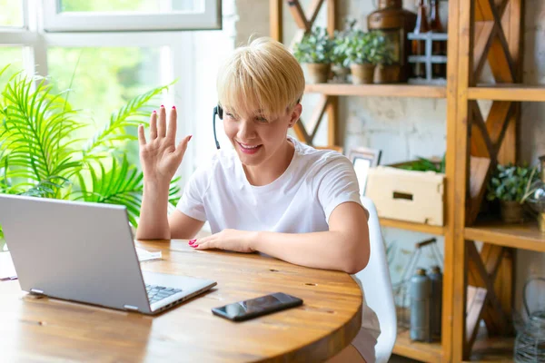 女性はノートパソコンを使って自宅で働いています。彼女はビジネスミーティング(オンラインビデオ通話)を主催しています。手を振って挨拶する. — ストック写真