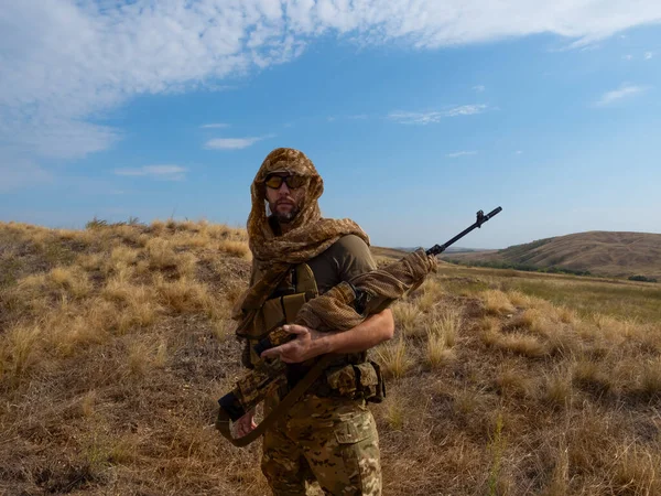 Francotirador-mercenario en ropa de camuflaje bajo el sol abrasador. Se para con un rifle y revisa la zona.. — Foto de Stock