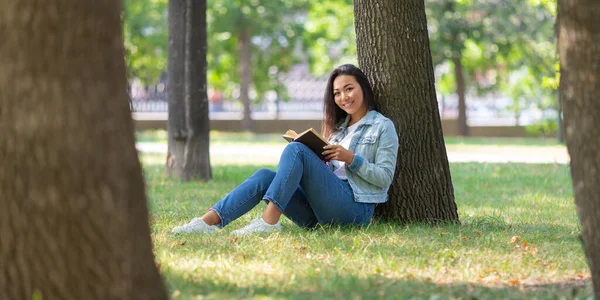 Azjatka z książką w parku. Siedzi na trawie z książką, patrzy w kamerę i uśmiecha się. Pojęcie odpoczynku od gadżetów, smartfonów i Internetu. Format zdjęcia 2x1. — Zdjęcie stockowe