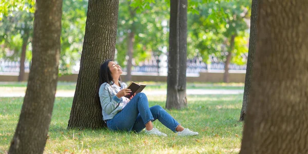 Azjatka z książką w parku. Siedzi na trawie w parku i patrzy obok siebie. Koncepcja detoksykacji cyfrowej. Format zdjęcia 2x1. — Zdjęcie stockowe