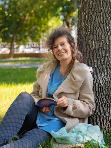 Kobieta w średnim wieku z książką w parku. Siedzi na trawie z książką, patrzy w kamerę i uśmiecha się. Pojęcie odpoczynku od gadżetów, smartfonów i Internetu. — Zdjęcie stockowe