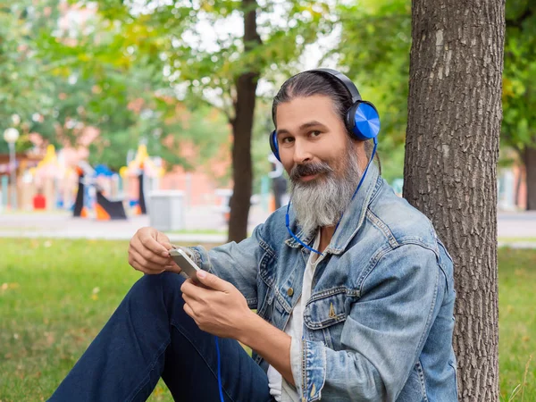Şehir parkında çimlerde otururken orta yaşlı bir adam müziğin tadını çıkarıyor. Kameraya bakıp gülümsüyor.. — Stok fotoğraf