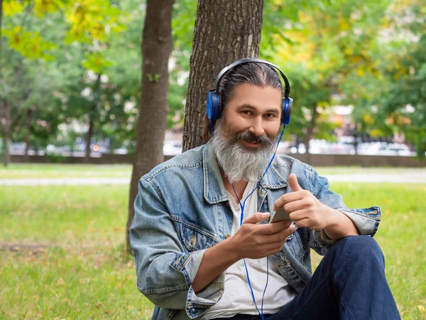 Şehir parkında çimlerin üzerinde otururken orta yaşlı bir adamın müziğin keyfini çıkardığı fotoğraf. Kameraya bakıp gülümsüyor.. — Stok fotoğraf