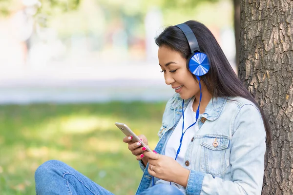 Femme asiatique dans les écouteurs avec smartphone. Elle sourit et écoute sa musique préférée ou sa leçon éducative. — Photo