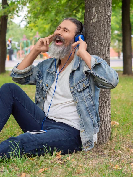 Şehir parkında çimlerin üzerinde otururken müziğin keyfini çıkaran orta yaşlı bir adamın fotoğrafı. Dikey fotoğraf.. — Stok fotoğraf