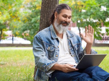 Çevrimiçi video görüşmesi sırasında orta yaşlı bir adam. Şehir parkındaki çimlerin üzerinde dizüstü bilgisayarla oturuyor. El sallıyor. Merhaba. Kesilmiş fotoğraf.