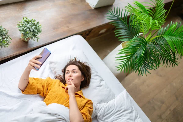 Cinta virtual (Virtual love): seorang wanita berbaring di tempat tidur dengan ponsel. Panggilan online untuk pacar virtual. Tampilan dari atas. — Stok Foto