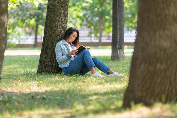 Azjatka czyta książkę w parku. Siedzi na trawie pod drzewem, czyta książkę i robi sobie przerwę od ciągłej pracy przy komputerze i stale jest online. — Zdjęcie stockowe