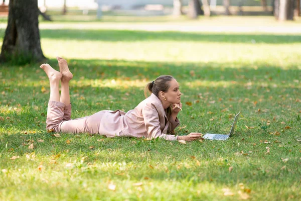 Zamyślona kobieta leży w parku na trawie przed monitorem laptopa. Koncepcja edukacji internetowej. — Zdjęcie stockowe