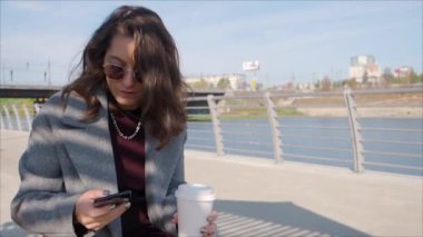 Bir fincan kahveyle dışarıda nehir kıyısında otururken akıllı telefonuna mesaj yazan bir kadın..