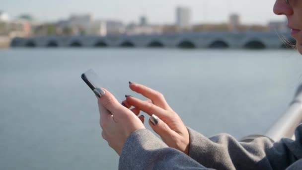 Şehir nehir kıyısında otururken akıllı telefonuyla mesaj yazan bir kadının yakın görüntüsü.. — Stok video
