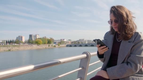 Donna digitando un messaggio di testo sul suo smartphone all'aperto mentre seduto sulla costa del fiume della città. — Video Stock