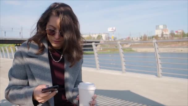 Γυναίκα με ένα φλιτζάνι καφέ πληκτρολογώντας μήνυμα κειμένου στο smartphone της εξωτερική, ενώ κάθεται στην ακτή του ποταμού της πόλης. — Αρχείο Βίντεο