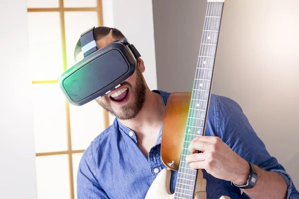 Adulto joven tocando la guitarra en casa usando el visor para virtual real — Foto de Stock