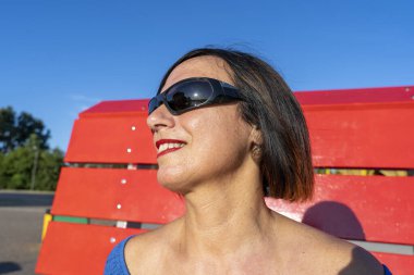 Mavi giyinmiş güzel bir kadın büyük, kırmızı bir bankta dinleniyor.