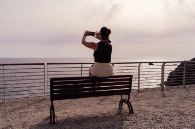 Güzel bir kadın gün batımında deniz kenarında bir bankta oturmuş fotoğraf çekiyor.