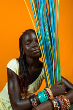 Gözleri kapalı, uzun renkli pipetlerle oynayan Afrikalı genç bir kız. Sarı arkaplanda stüdyo çekimi 