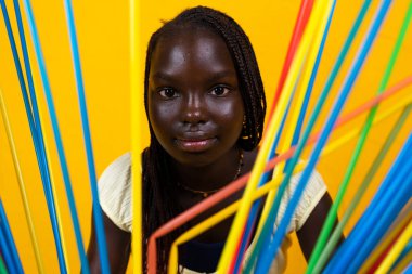 Uzun renkli pipetlerin arkasındaki Afrikalı genç kızın portresi. Sarı arkaplanda stüdyo çekimi 