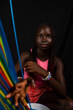 Genç ve güzel Afrikalı bir gencin portresi uzun renkli pipetlerin arkasında duruyor. Siyah arkaplanda stüdyo çekimi