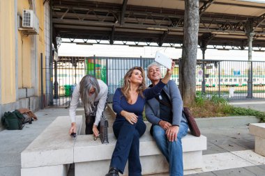 Bir grup yetişkin, tren istasyonunun önündeki beton bir bankta otururken akıllı telefonlarıyla selfie çekiyor.
