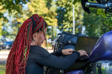 Motosikletinin üzerinde otururken dizüstü bilgisayarda yazan renkli rastalı bir kadın bisikletçi.